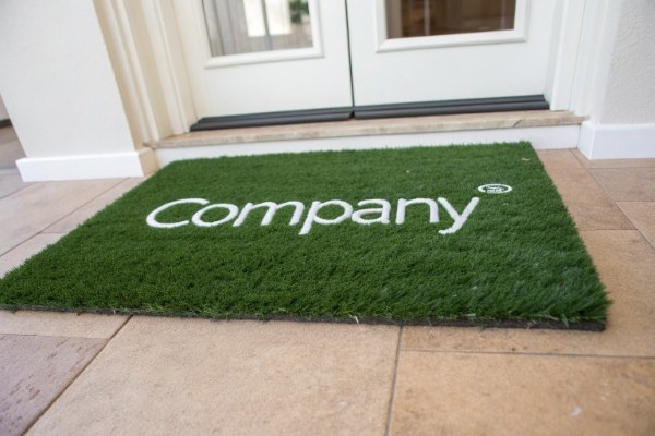A custom artificial grass doormat with a company's white logo woven into the green turf